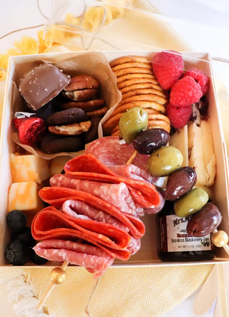 all of the goodies inside the diy individual charcuterie box with meat and olives on skewers, nuts and dried fruit in bakers cup, fruit, cracker and cheese scattered around.