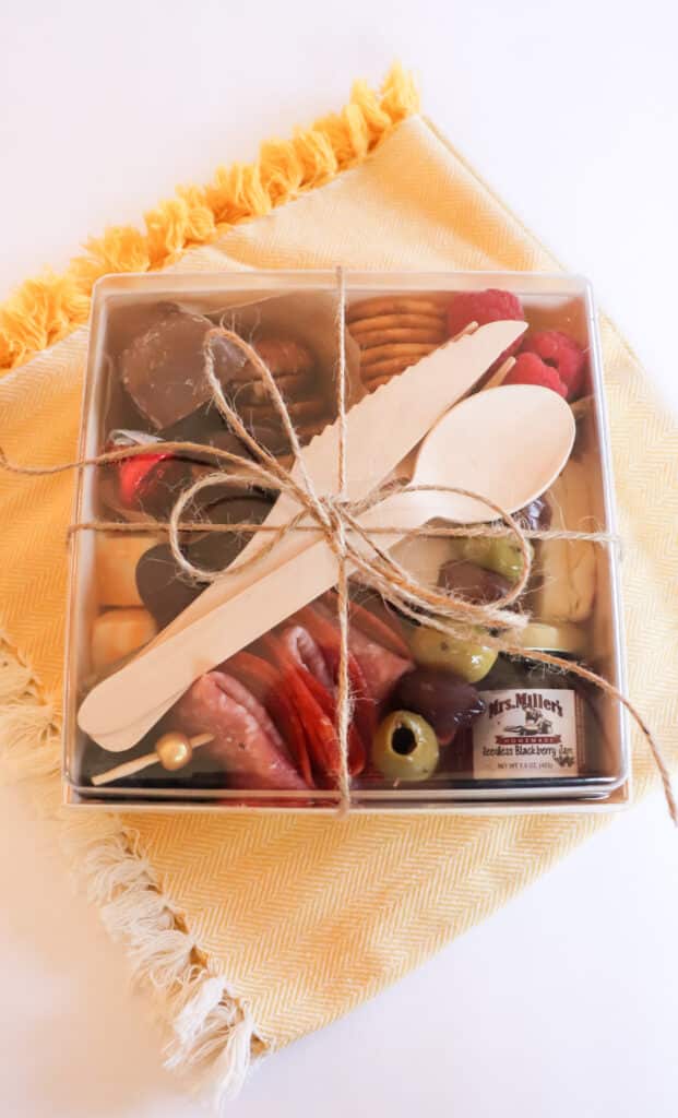 charcuterie box with meat, cheese, fruit, and silverware tied with twine sitting on a yellow napkin.