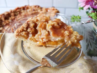 Slice of peach pie on a plate with the rest of the pie in the background.