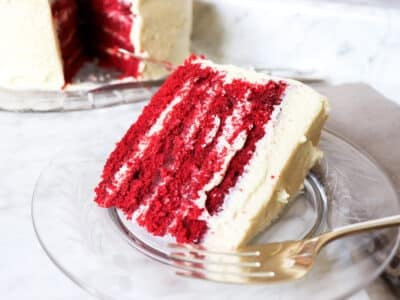 Slice of red velvet cake on a plate with the whole cake sitting behind it.