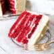 Slice of red velvet cake on a plate with the whole cake sitting behind it.