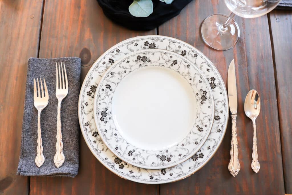 One place setting of china with a salad plate sitting on a dinner plate, a cloth napkin, and silver flatware.