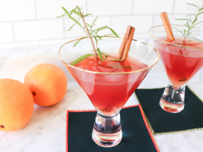Two Cosmos in glasses sitting on the counter with two oranges in the background.