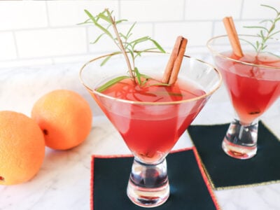 Two cocktails with rosemary and cinnamon garnishes and oranges in the background.
