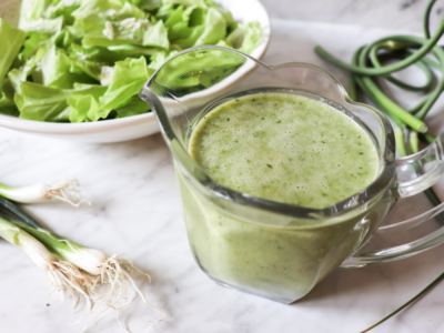 Garlic scape dressing in a clear pitcher with lettuce in a bowl and garlic scapes and onions on the counter.