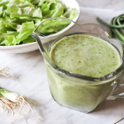 Garlic scape dressing in a clear pitcher with lettuce in a bowl and garlic scapes and onions on the counter.