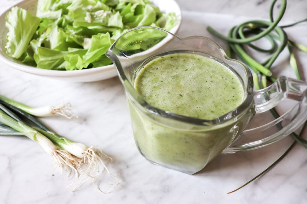 Garlic scape dressing in a clear pitcher with lettuce in a bowl and garlic scapes and onions on the counter.
