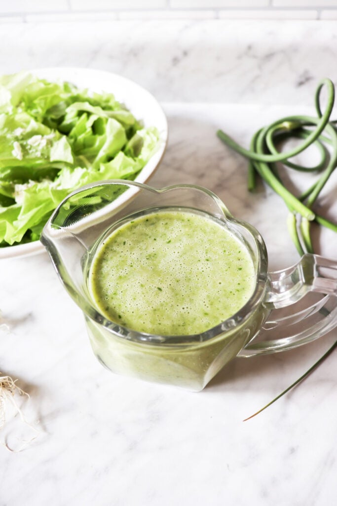 Garlic scape dressing in a clear pitcher with lettuce in a bowl and garlic scapes and onions on the counter.