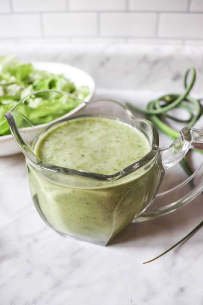 Garlic scape dressing in a clear pitcher with lettuce in a bowl and garlic scapes and onions on the counter.