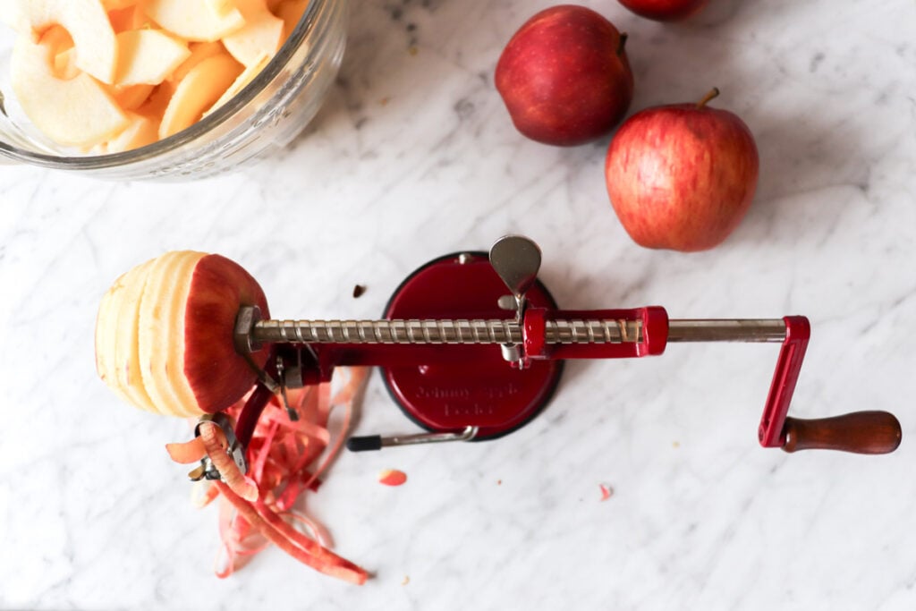 Apple peeler, corer, slicer on the counter with apples lying around.