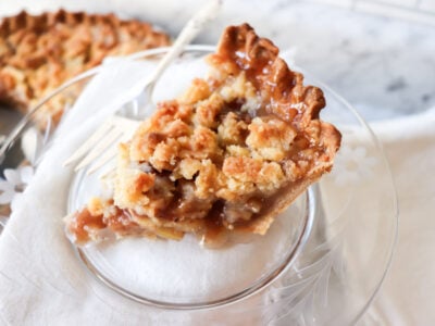 Slice of apple pie on a plate with the rest of the pie in the background.