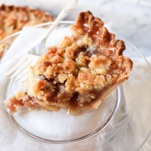 Slice of apple pie on a plate with the rest of the pie in the background.