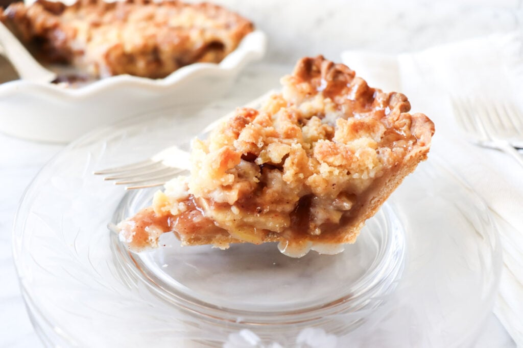 A slice of apple pie on a plate with the rest of the pie in the backgound.